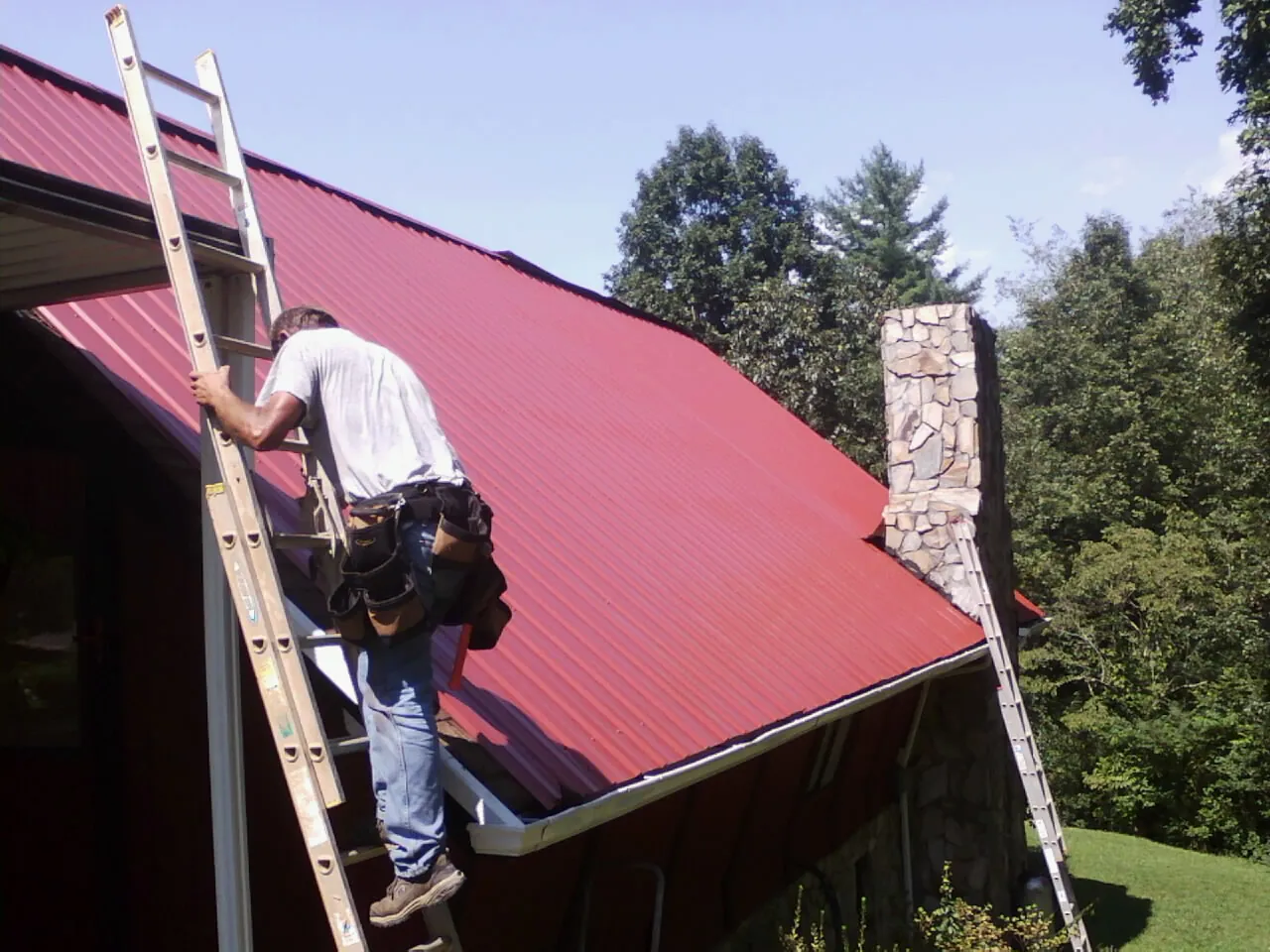 Roofer on a ladder beside a red metal roof and stone chimney — Soffit & Fascia Repair by Roofing Brothers in Harrah